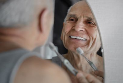 Un homme âgé souriant dans le miroir tout en tenant une brosse à dents.