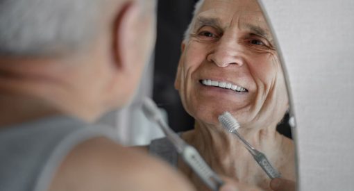 Un homme âgé souriant dans le miroir tout en tenant une brosse à dents.
