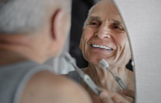 Un homme âgé souriant dans le miroir tout en tenant une brosse à dents.
