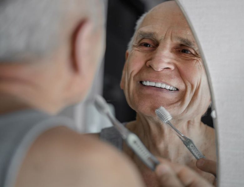 Un homme âgé souriant dans le miroir tout en tenant une brosse à dents.